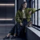 A seated fashion model in a green bomber jacket, formed from code characters, beside a window.
