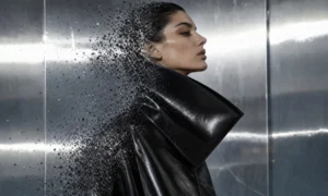 Illustration of a model in a black coat dissolving into particles against a steel backdrop.
